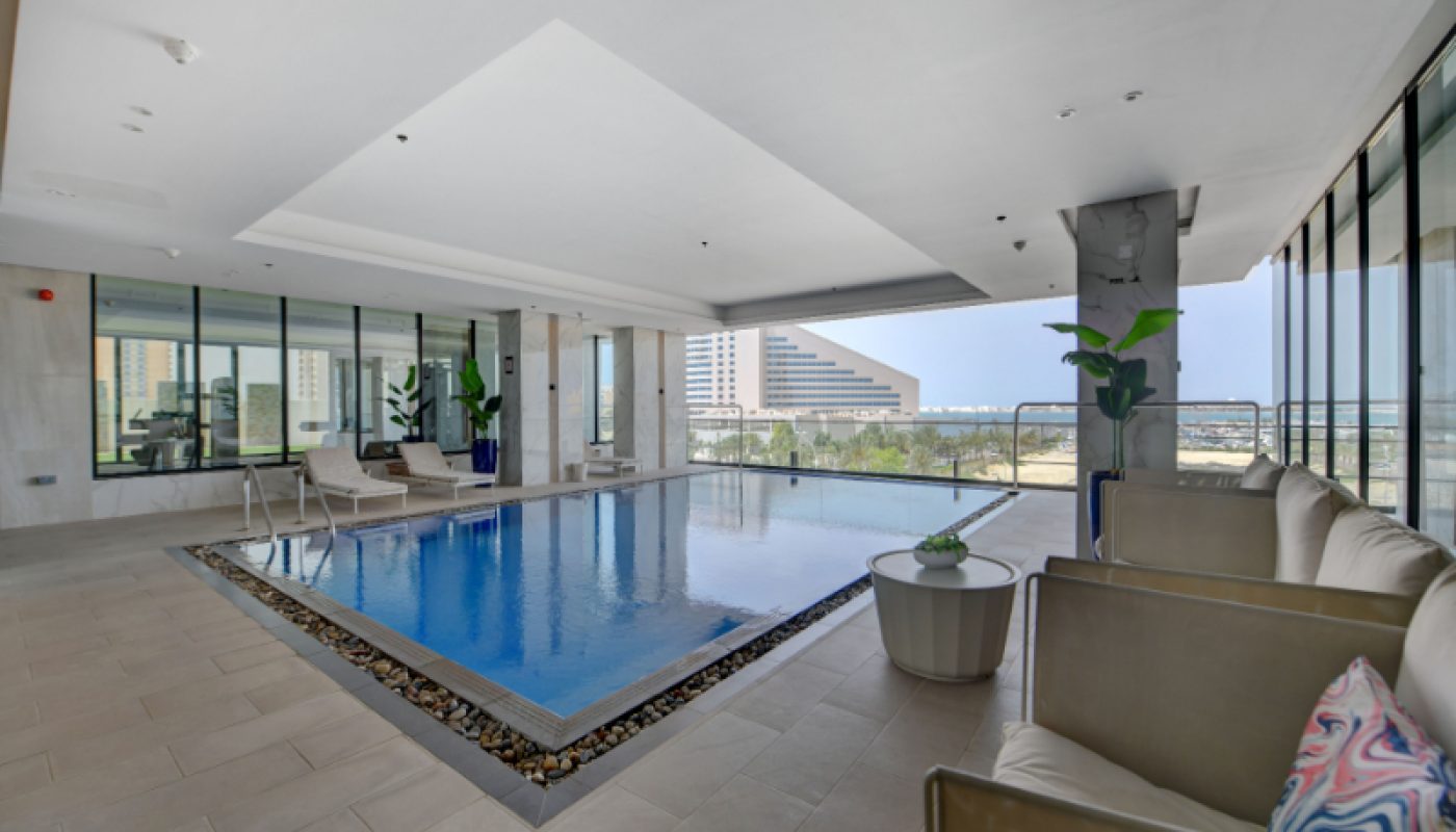 Modern indoor pool area with tile flooring, lounge chairs, potted plants, and large windows providing a view of city buildings outside.