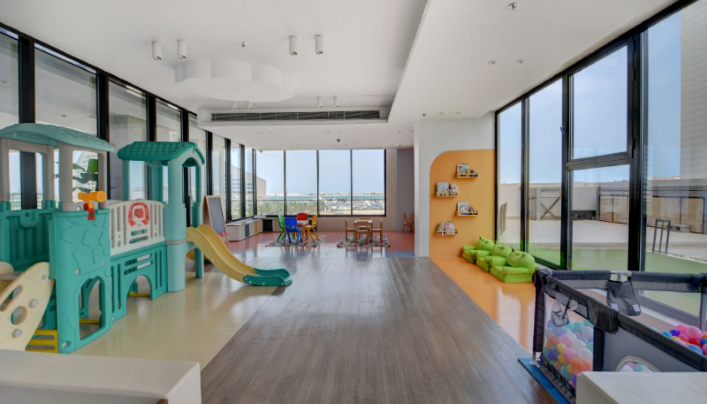 Spacious children's playroom with large windows, a play structure with slides, a ball pit, bean bags, and tables with chairs, overlooking a city view.