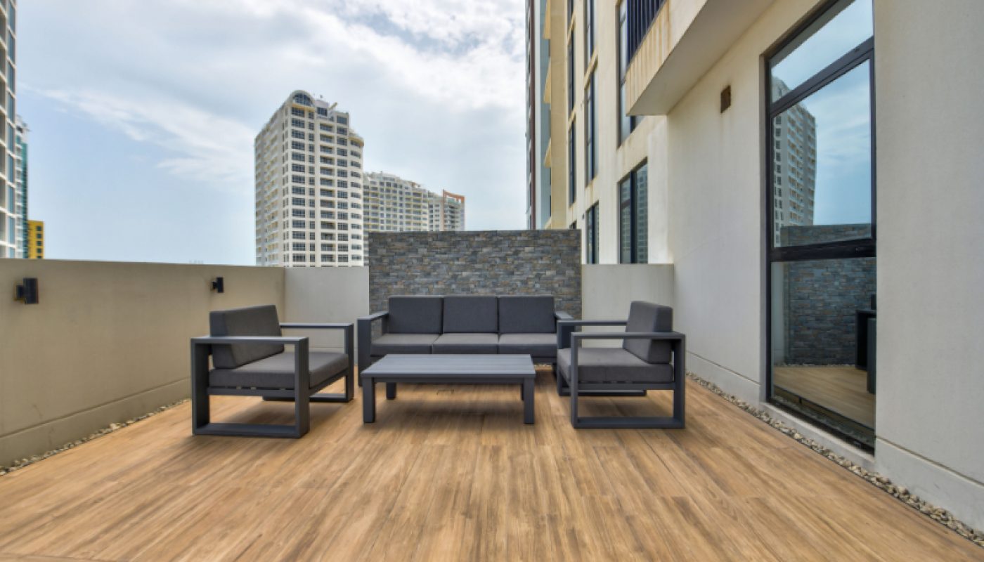 Modern outdoor patio with wooden flooring, a gray sofa, two armchairs, a coffee table, and surrounding tall buildings under a partly cloudy sky.