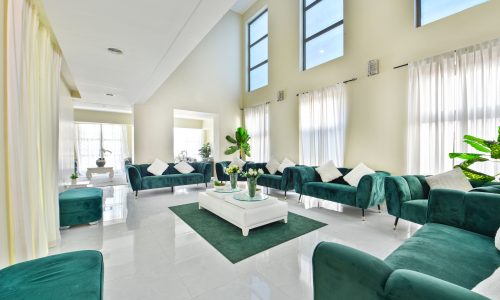 Bright and spacious living room in an amazing 5BR villa for rent on Tala Island, featuring white walls and high ceilings, furnished with green velvet sofas and chairs, and complemented by white curtains