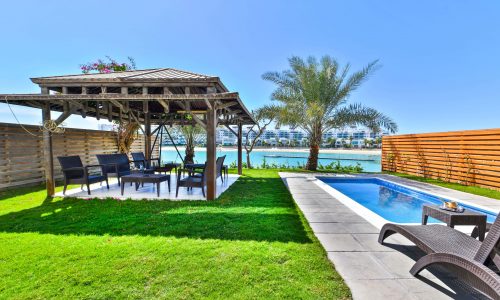 Backyard of a 5BR Villa on Tala Island with a pool, pergola, and seating area overlooking a water body on a sunny day.