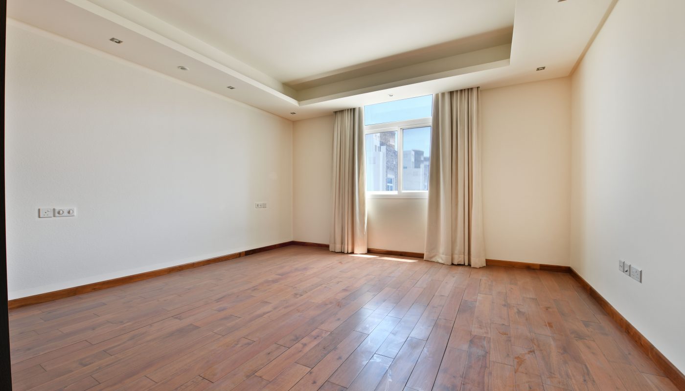A bright, empty room with wooden flooring, beige walls, recessed lighting on the ceiling, and a window with beige curtains allowing natural light to enter in this luxurious villa for sale in Janabiyah.