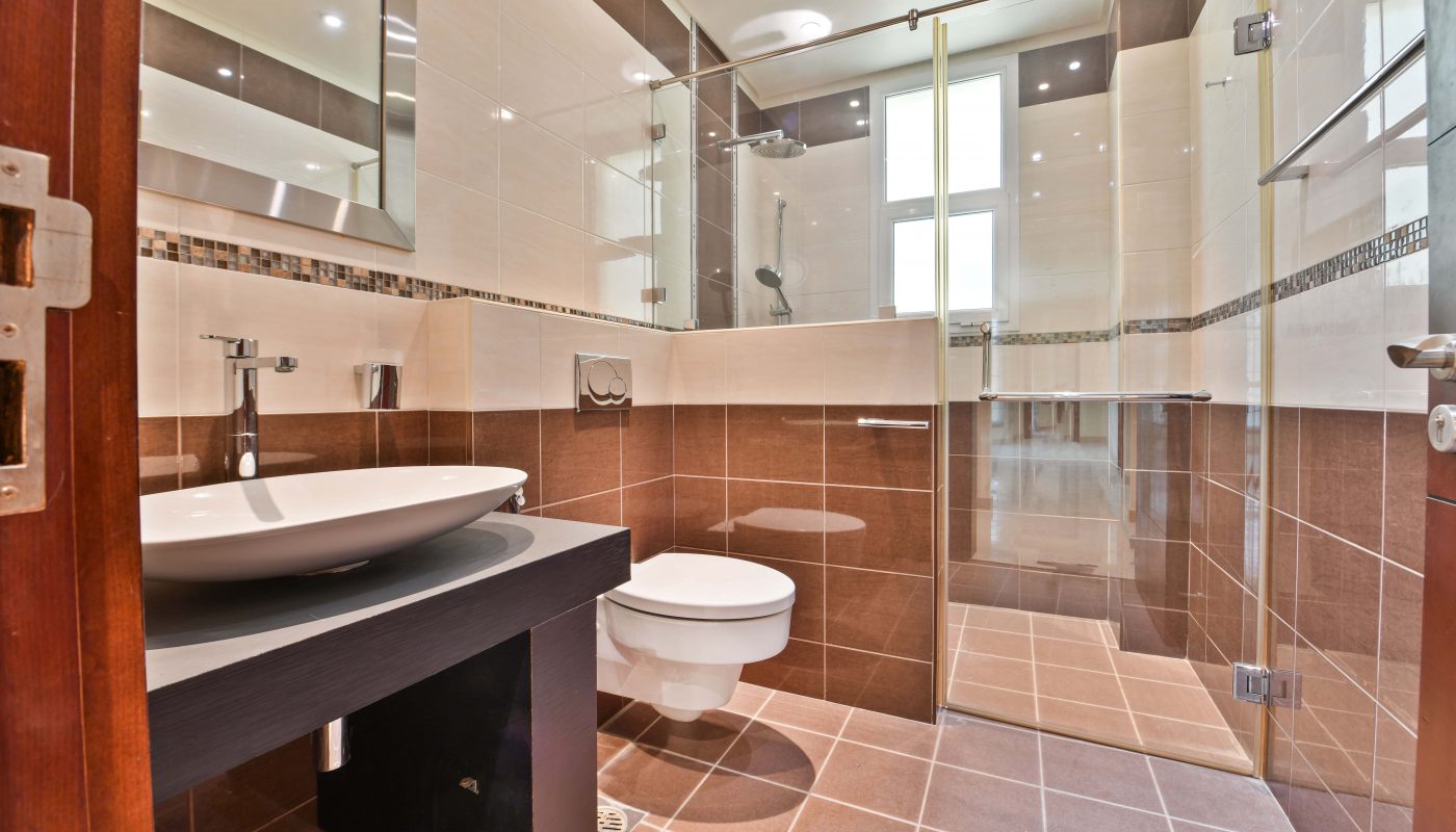 A modern bathroom in a luxurious villa for sale in the Janabiyah area, complete with a glass-enclosed shower, a wall-mounted toilet, and a sink with a round basin. The room features brown and beige tile walls and floors, accented by recessed ceiling lights.