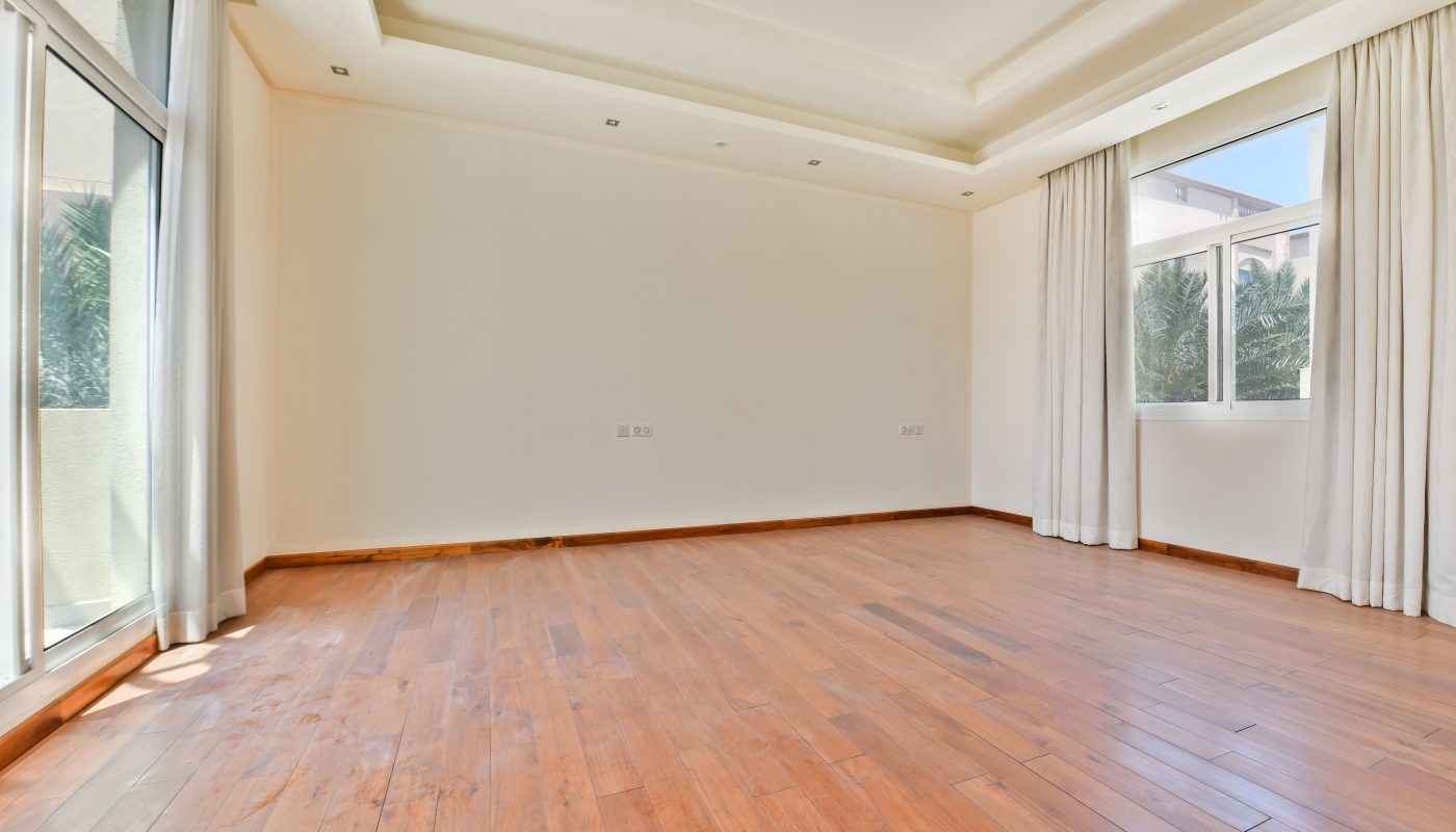 An empty room with wooden flooring, large windows with white curtains in a luxurious villa for sale. The walls are plain white with recessed lighting on the ceiling, and trees are visible through the windows. Experience serene living in the sought-after Janabiyah Area.