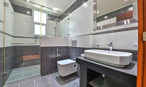 Modern bathroom in a luxurious villa in Janabiyah featuring a glass-enclosed shower, wall-mounted toilet, rectangular sink on a black countertop, large mirror, and gray and white tiled walls and floor.