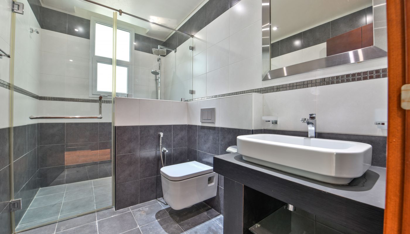 Modern bathroom in a luxurious villa in Janabiyah featuring a glass-enclosed shower, wall-mounted toilet, rectangular sink on a black countertop, large mirror, and gray and white tiled walls and floor.