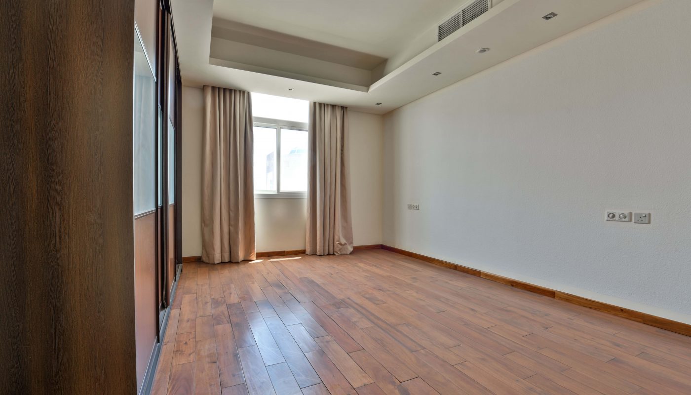 This luxurious villa for sale in Janabiyah features an empty room with wooden flooring, a large window adorned with beige curtains, recessed ceiling lights, and pristine white walls. A built-in cabinet with mirrored doors graces the left side, enhancing the elegant ambiance.