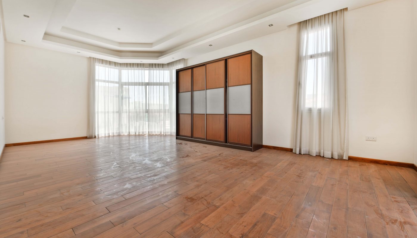 An empty room in a luxurious villa for sale in Janabiyah, featuring wooden flooring, large windows with white curtains, and a wooden wardrobe against the wall. The room boasts a tray ceiling with recessed lighting.