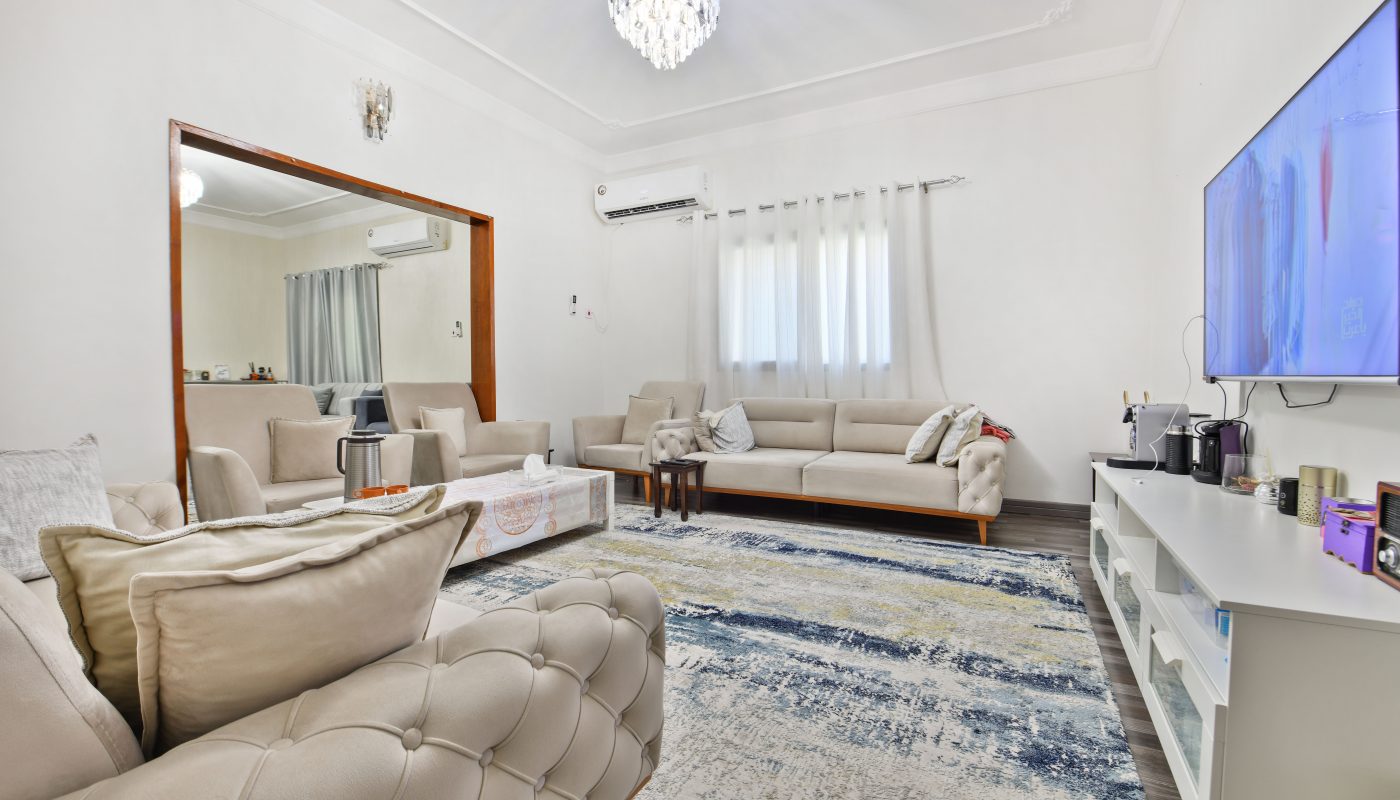 Modern living room in a Riffa villa for sale, featuring beige sofas, a large blue patterned rug, a TV screen displaying blue graphics, and a crystal chandelier.