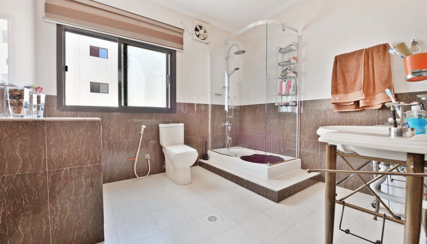 Modern bathroom interior in a villa for sale, featuring a small bathtub, white toilet, and sink, complemented by dark brown tiles and a frosted glass window.