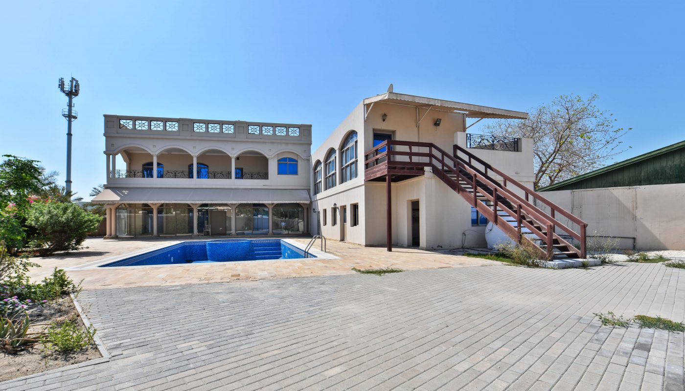 A two-story house with a balcony and an external staircase, featuring a swimming pool in the front yard, surrounded by paved walkways and some greenery. Auto Draft