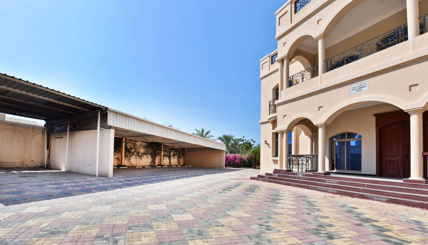 A large residential building with a beige exterior, multiple balconies, and an auto draft parking area, situated under a clear blue sky.