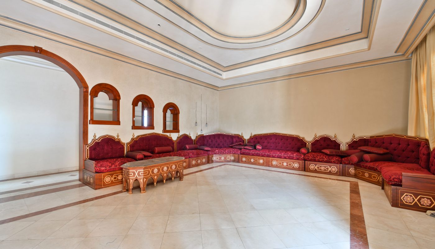 Spacious, elegant room with ornate red sofas, arched windows, and a bright, marbled floor under a decorative ceiling.