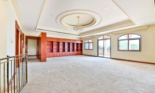 Spacious empty room in a Janusan villa with carpeted floors, large windows, a chandelier, and built-in red shelving unit.