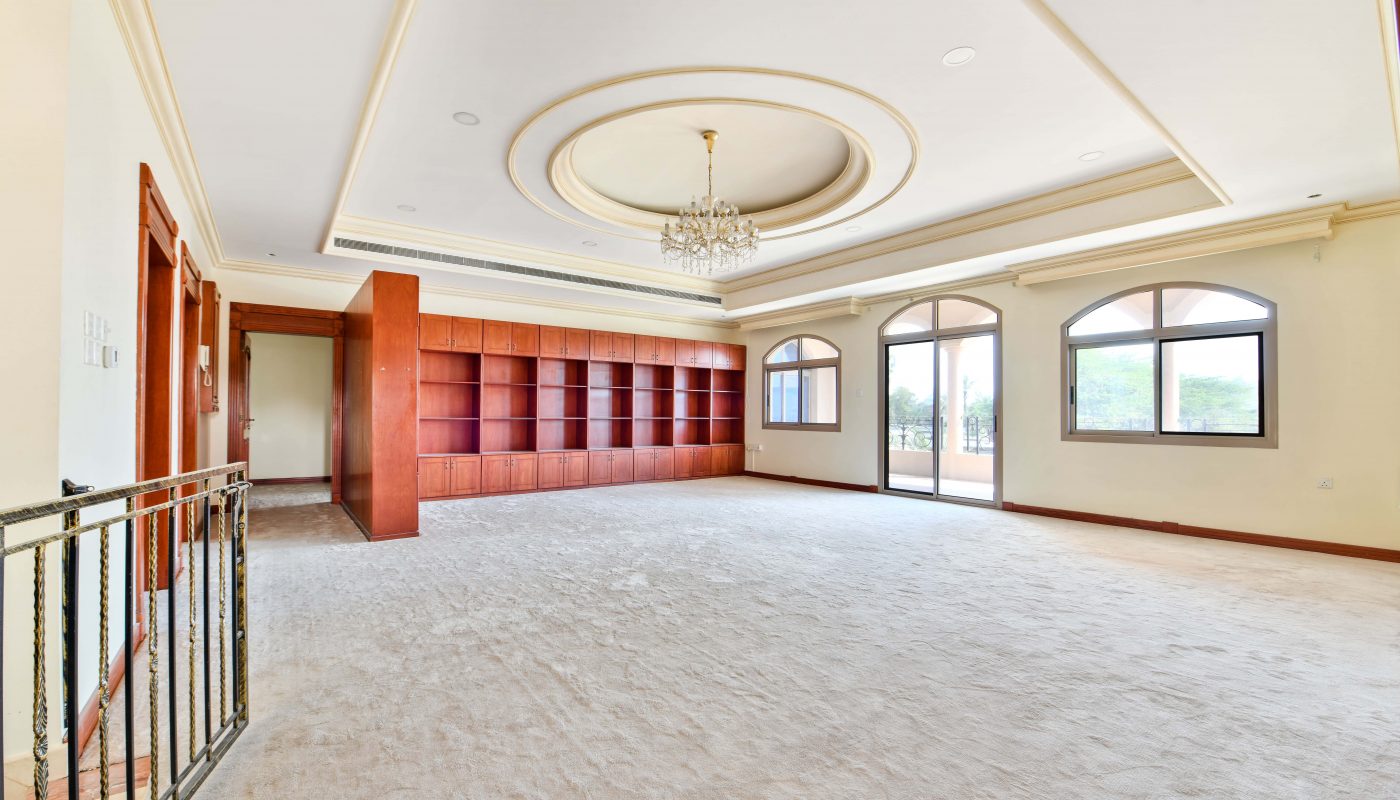 Spacious empty room in a Janusan villa with carpeted floors, large windows, a chandelier, and built-in red shelving unit.