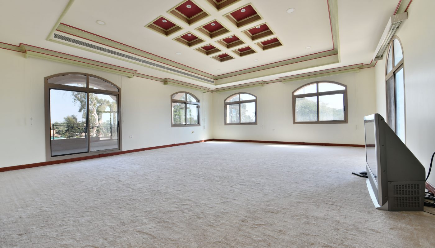 Empty room in a spacious villa with beige carpet, white walls with green trim, a decorative red and green ceiling, large windows and a glass door, and a TV on the floor.