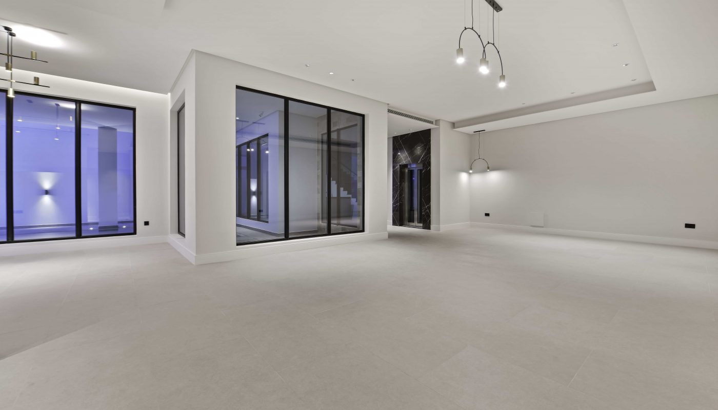 Spacious empty modern room with large windows, light-colored tiled floor, white walls, and contemporary ceiling lights.
