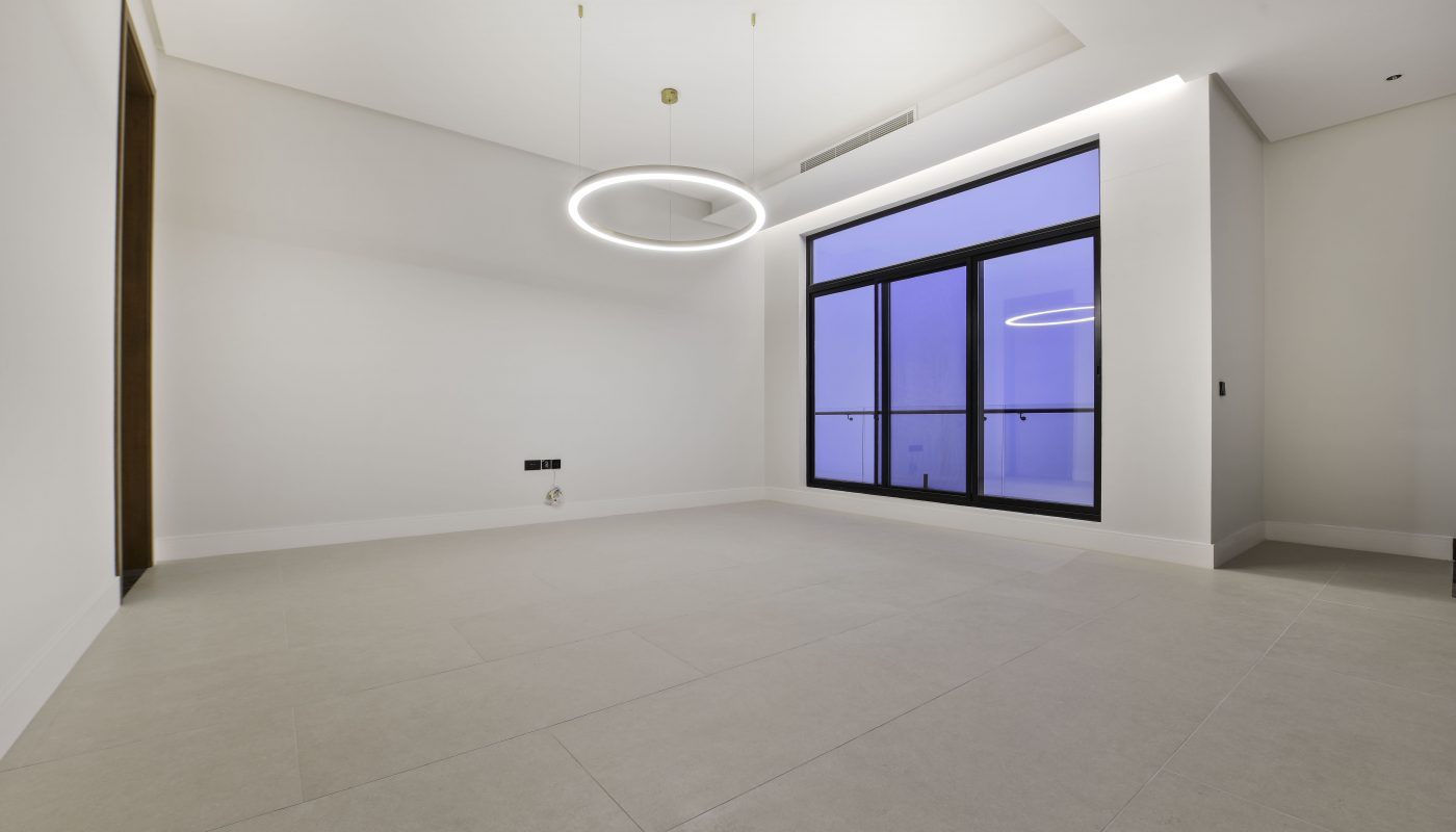 Empty modern room with large floor tiles, a circular ceiling light, white walls, and a window with a blue-tinted view.