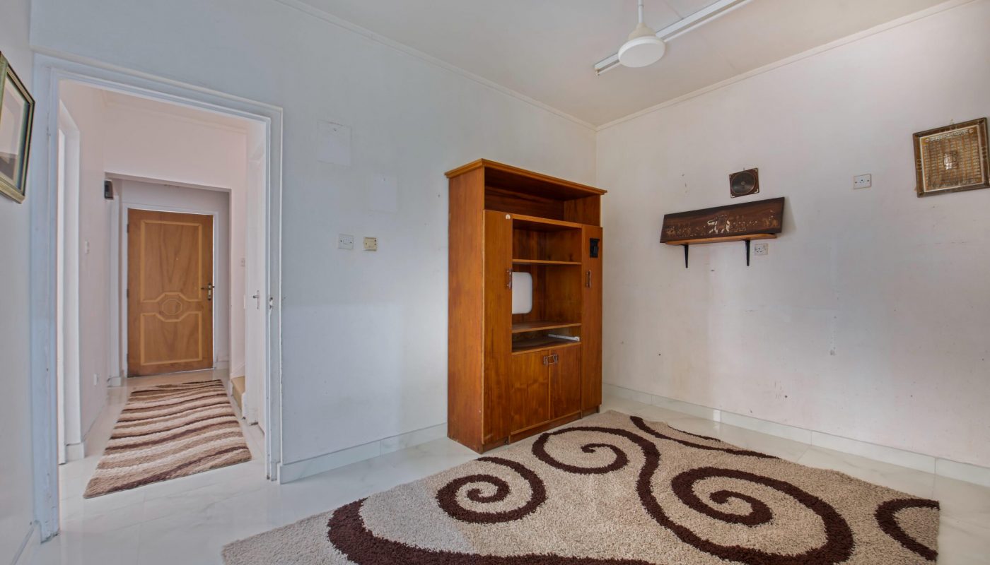 A sparsely furnished room with a ceiling fan, a wooden cabinet, light-colored walls, and a patterned rug. An open doorway leads to a hallway with another door visible.