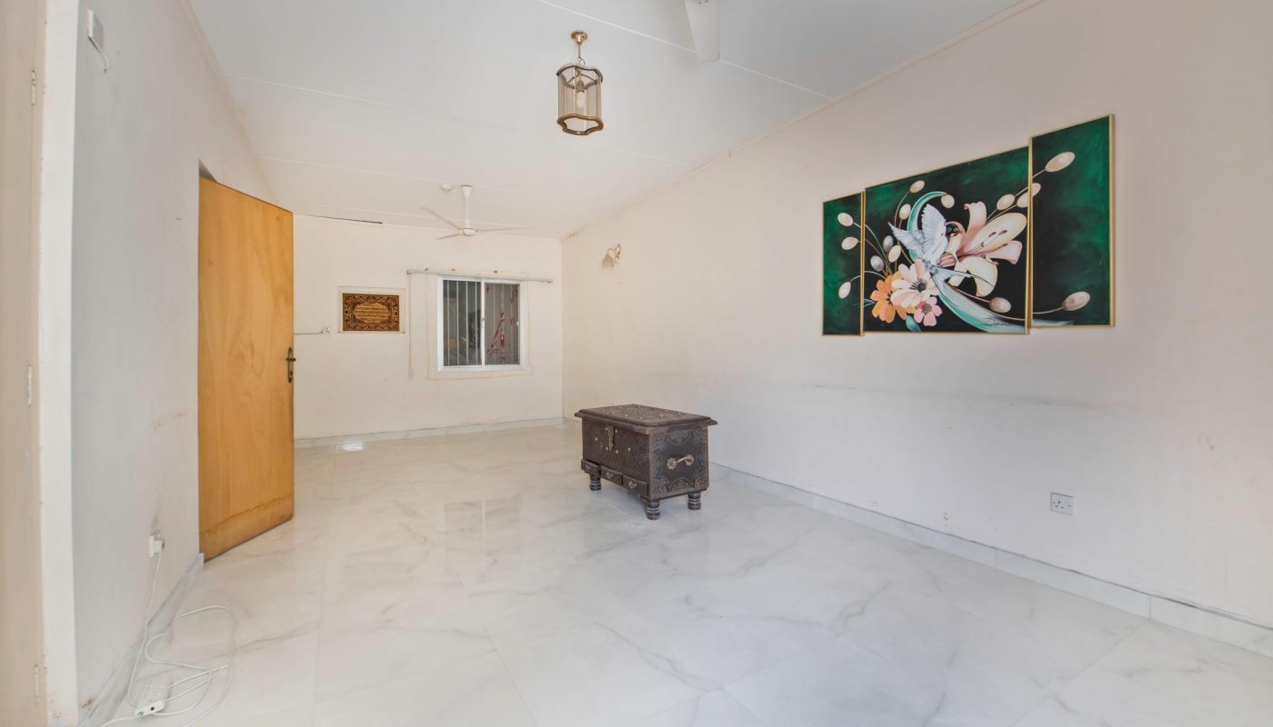 A sparsely furnished room with white tiled floor, a wooden chest in the center, a floral painting on the wall, ceiling fans, and a window with bars.