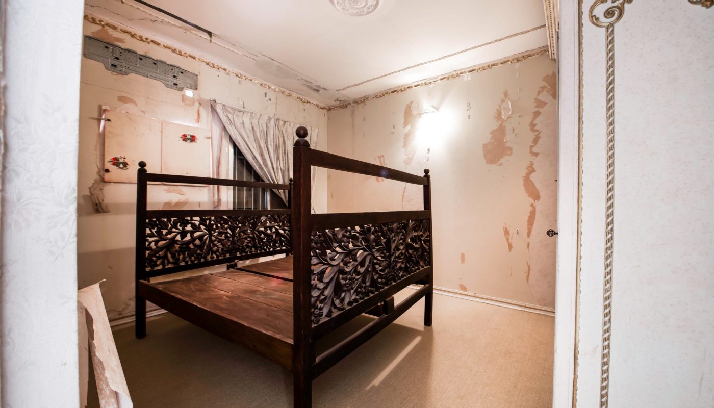 A wooden bed frame stands in a room with peeling wallpaper, exposed wall patches, a covered window, and minimal furnishings.