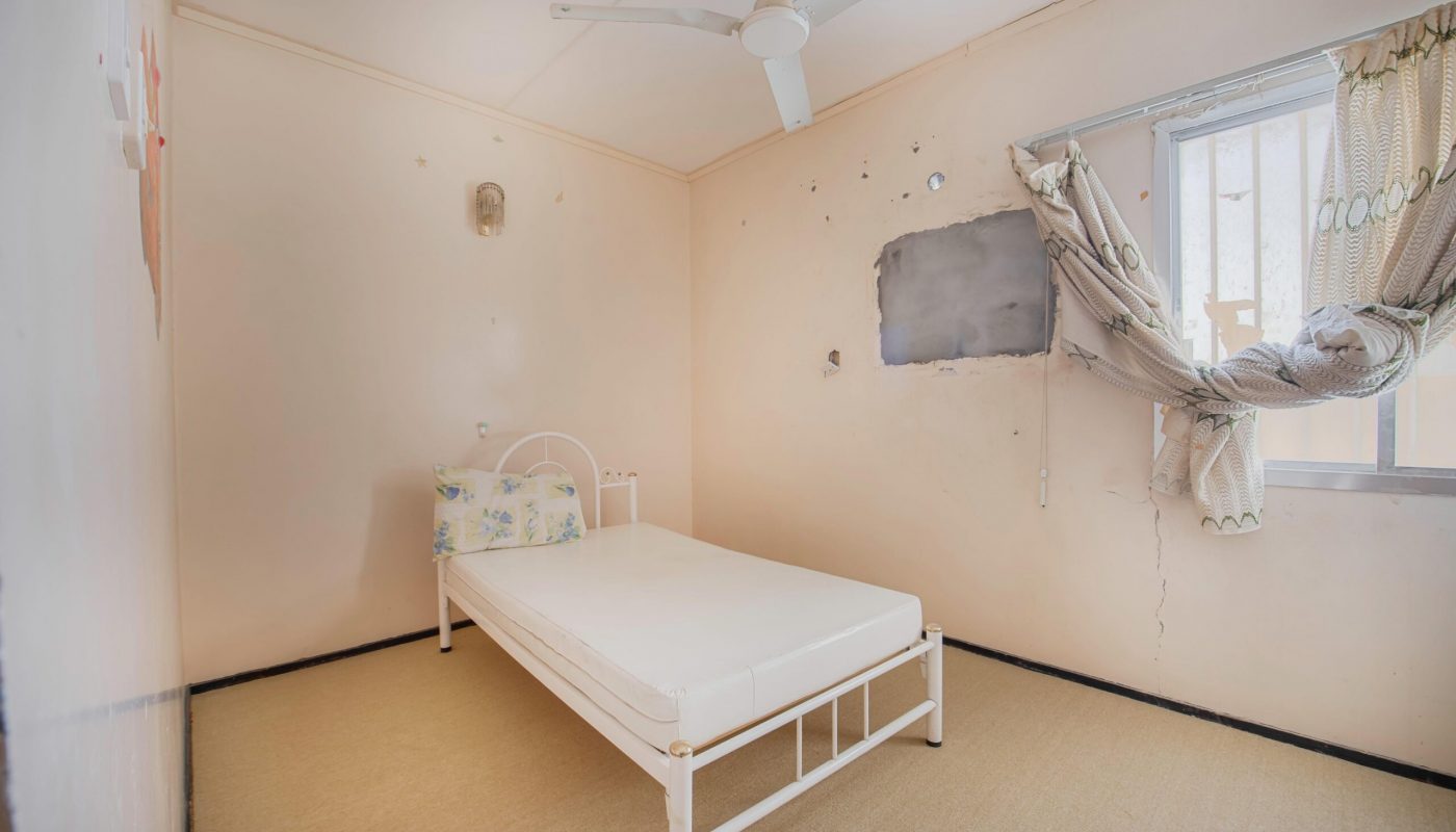 A small, sparse bedroom with a single metal bed, bare mattress, damaged walls, and a curtain hanging loosely from a window.
