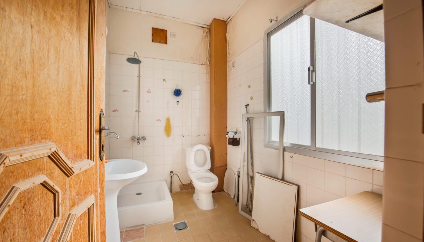 A small bathroom with a toilet, sink, shower, tiled walls, frosted window, and miscellaneous items leaning against the wall.