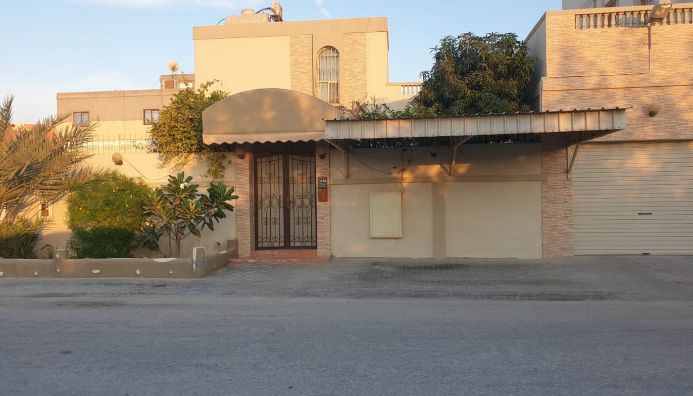 A beige two-story house with a gated entrance, large awning, and greenery in front, situated along a quiet street under a clear sky.