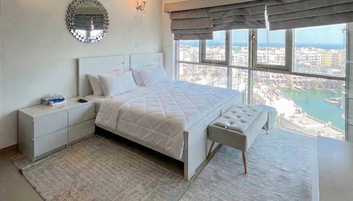 Modern bedroom with a large bed, two nightstands, a bench, rugs, wall mirror, and large windows overlooking a marina with boats.