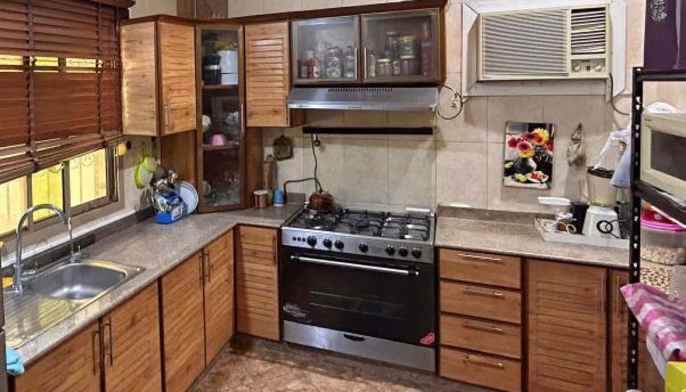 A tidy kitchen with wooden cabinets, a gas stove, a stainless steel sink, an air conditioner, a ceiling fan, and tiled floors and walls.