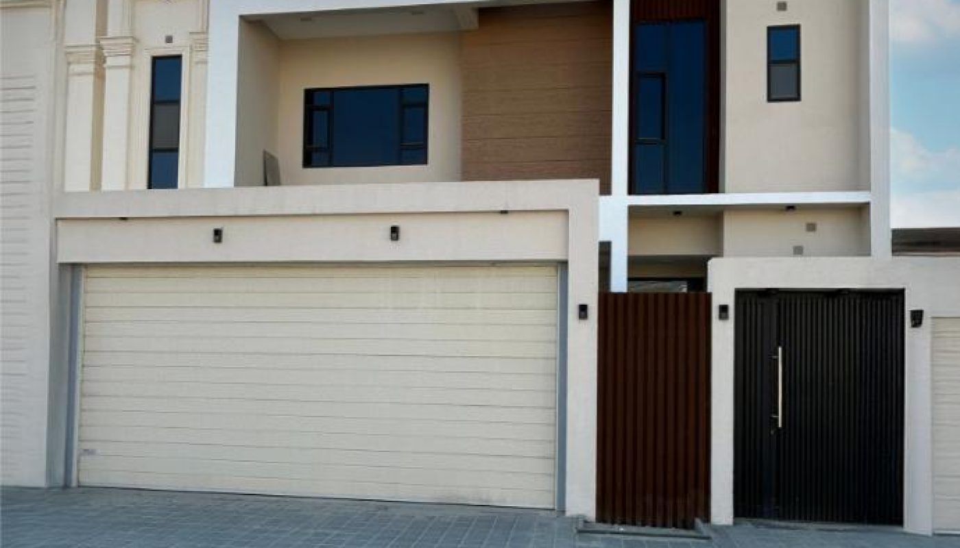 Modern two-story house with flat roof, beige exterior, large garage doors, vertical black accents, and multiple rectangular windows, set against a partly cloudy sky.
