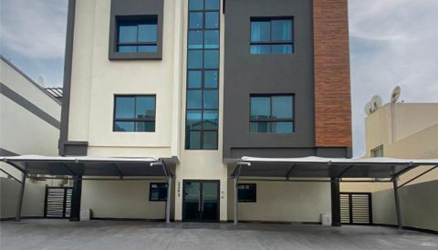 Modern three-story apartment building with a geometric facade, large windows, and covered parking spaces in front, set under a partly cloudy sky.