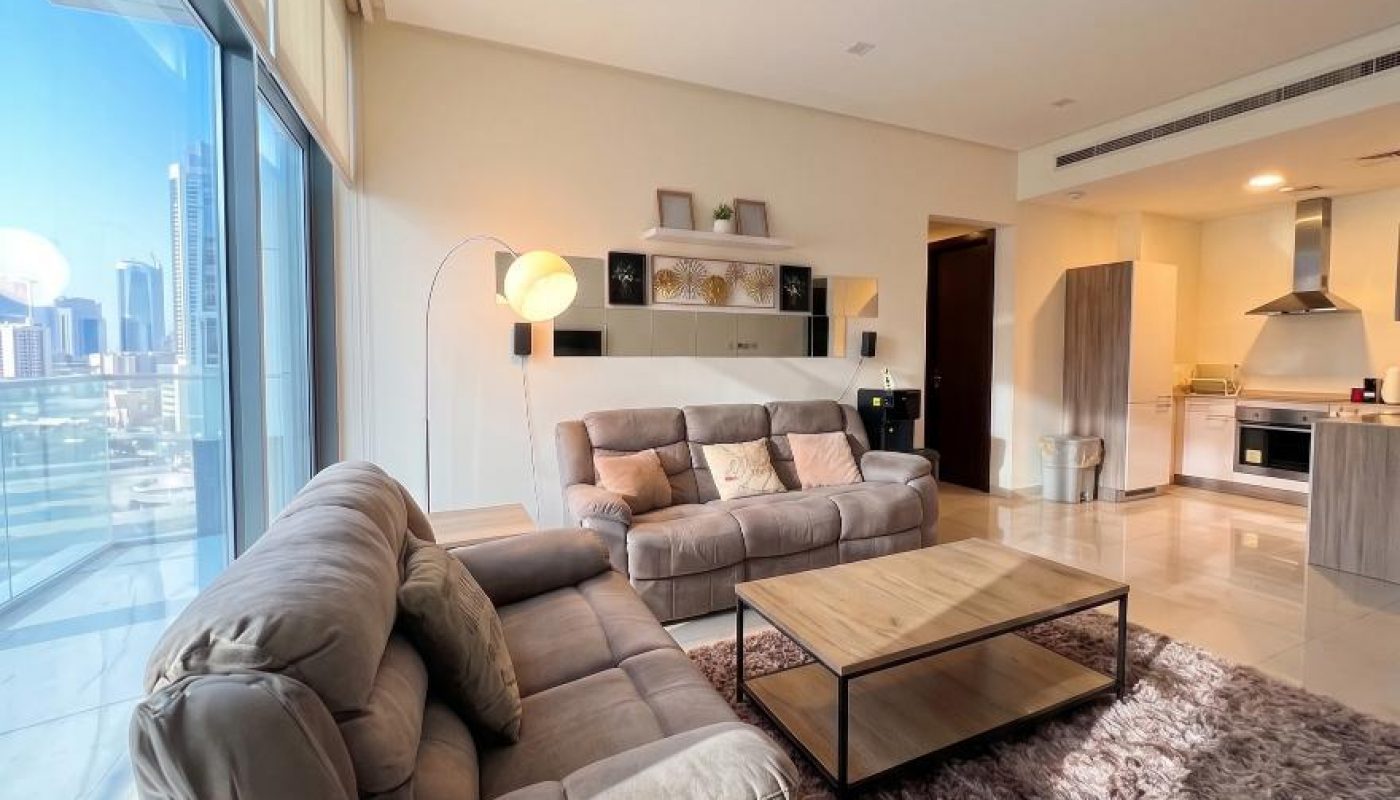 Modern living room with two gray sofas, a coffee table, a rug, and large windows overlooking a city skyline; kitchen area visible in the background.