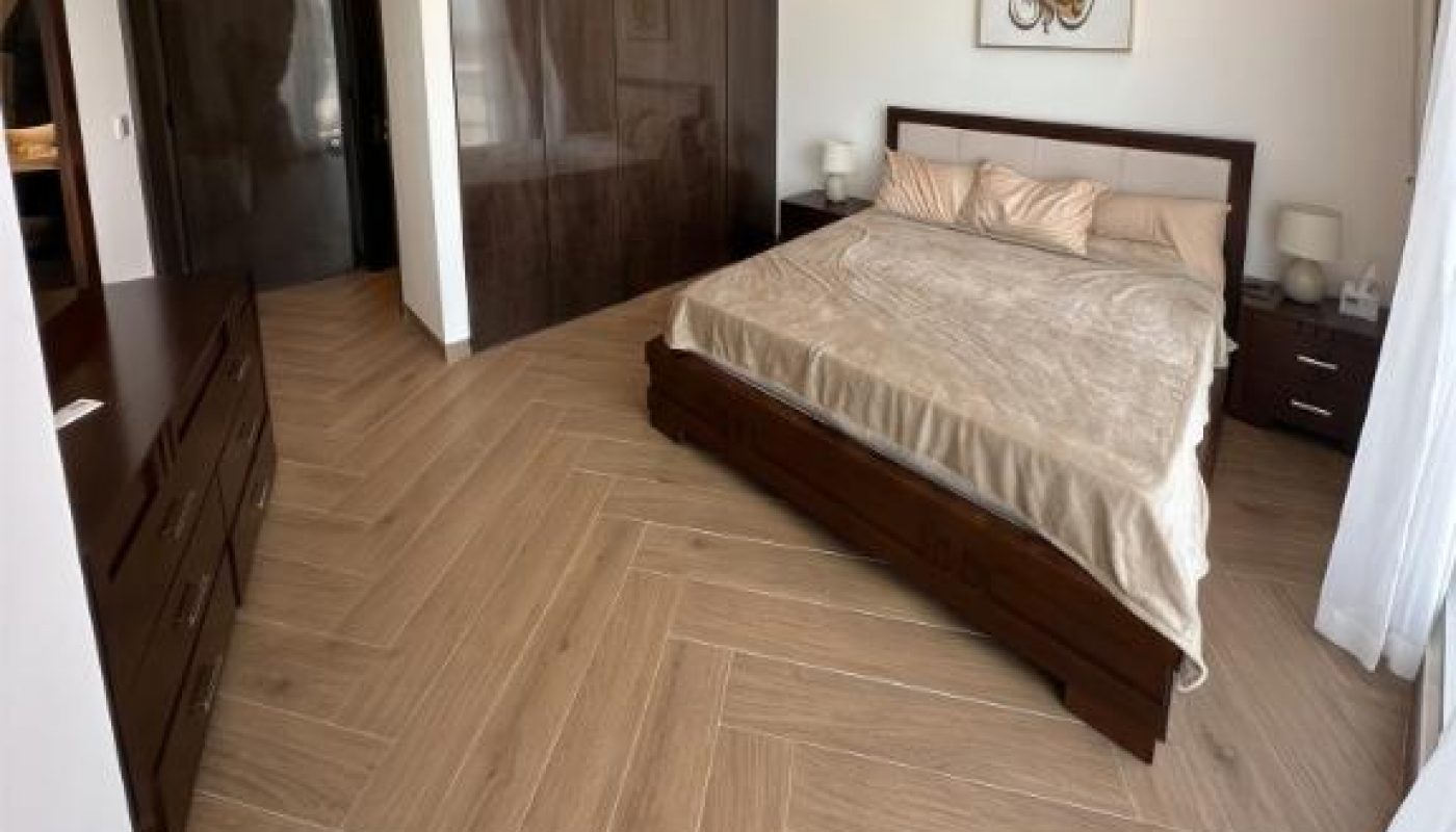 Modern bedroom with a large bed, wood furniture, and herringbone-patterned wood floor, featuring a large window with sheer curtains and neutral decor tones.