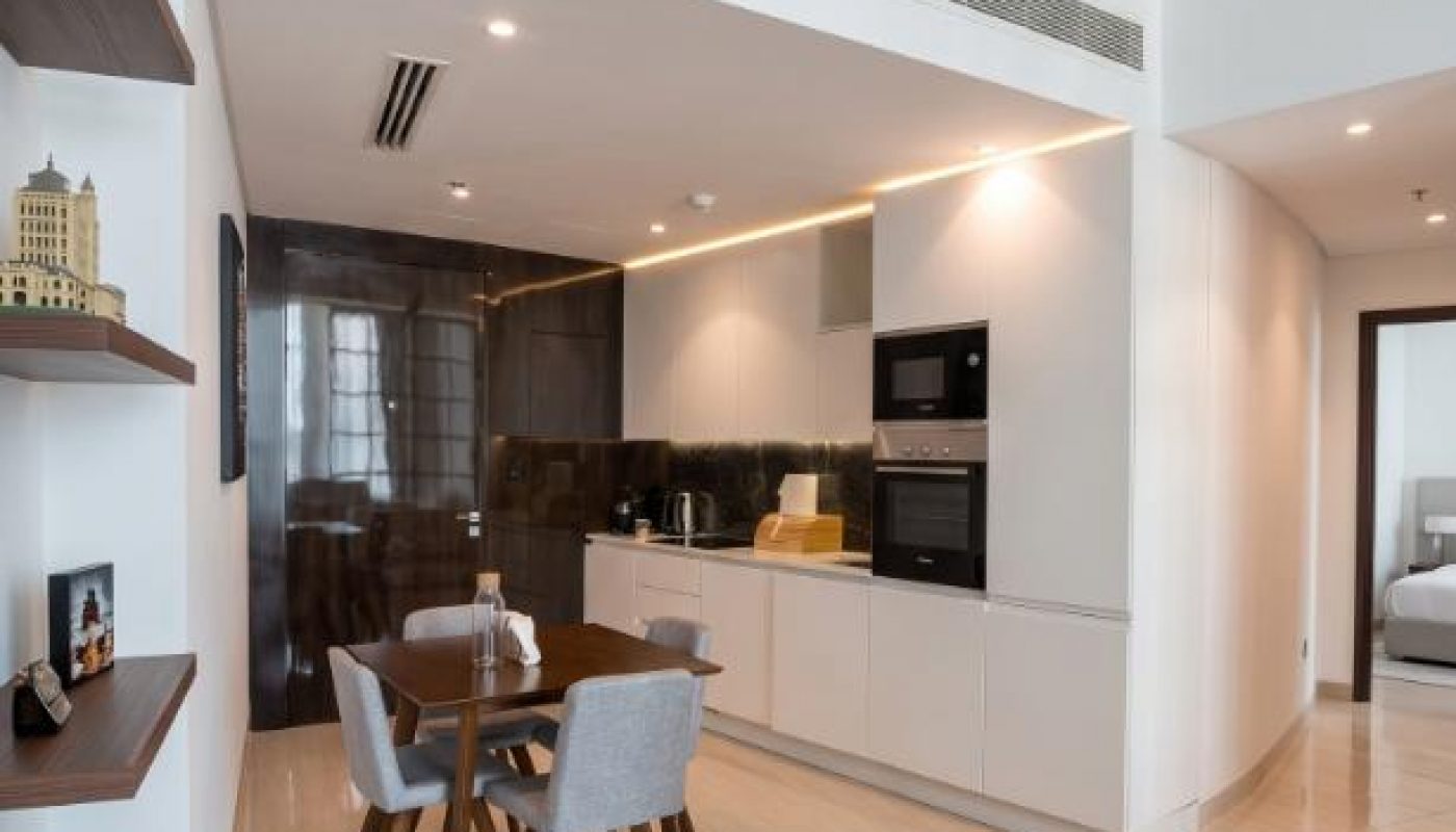 Modern kitchen and dining area with white cabinets, wooden table and four chairs, open shelving on the left, and a doorway leading to another room.