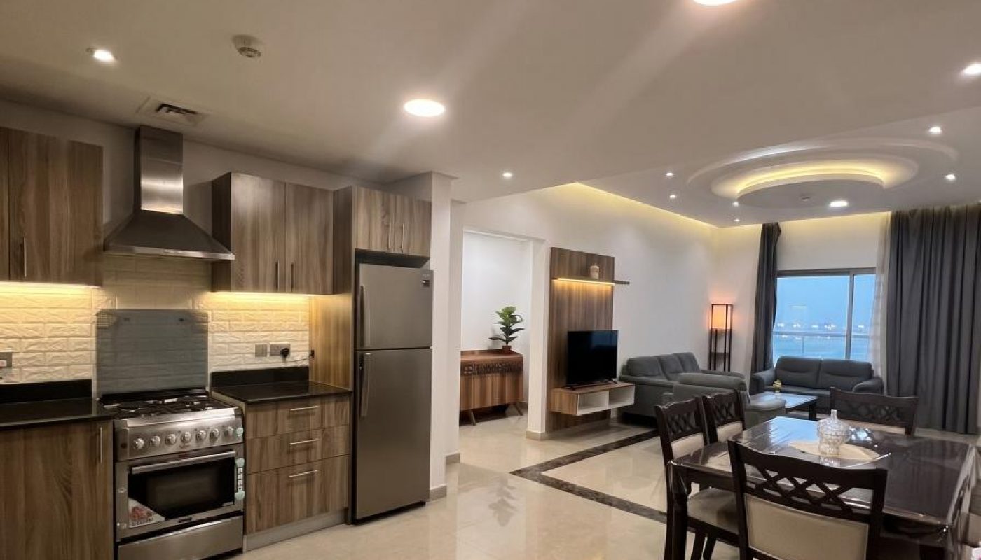 Modern open-plan kitchen, dining, and living area with wood cabinetry, stainless steel appliances, tiled flooring, neutral colors, and a large window with city or waterfront view.