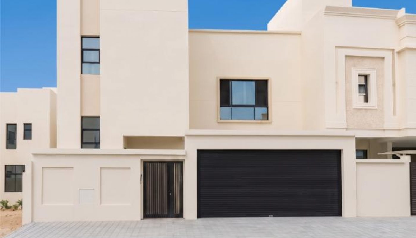 Modern beige two-story house with a black garage door and paved driveway under a clear blue sky.