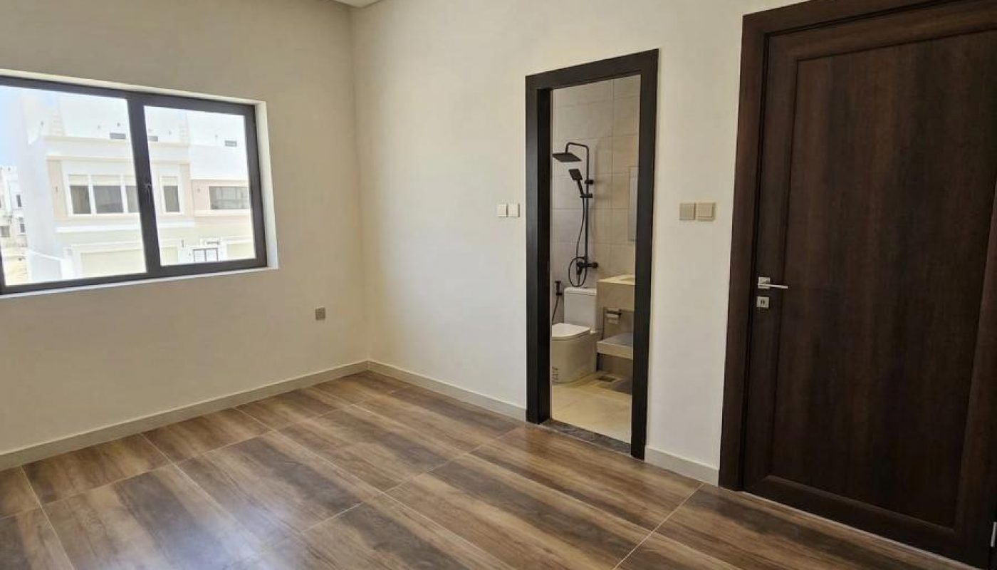 Empty room with wood-patterned floor, white walls, a ceiling fan, large window, a dark wooden door, and an open door leading to a bathroom with a shower visible.