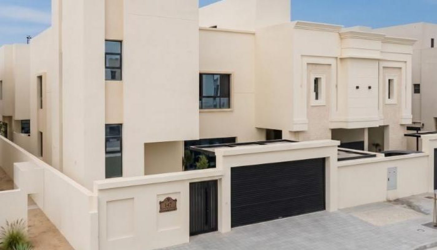 Modern two-story beige house with flat roof, black garage door, and minimal landscaping, situated on a paved street under a clear blue sky.