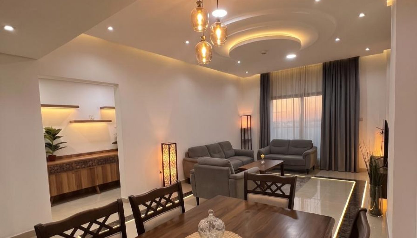 Modern living and dining area with wooden dining table, grey sofas, warm lighting, decorative ceiling fixtures, and floor-to-ceiling curtains.