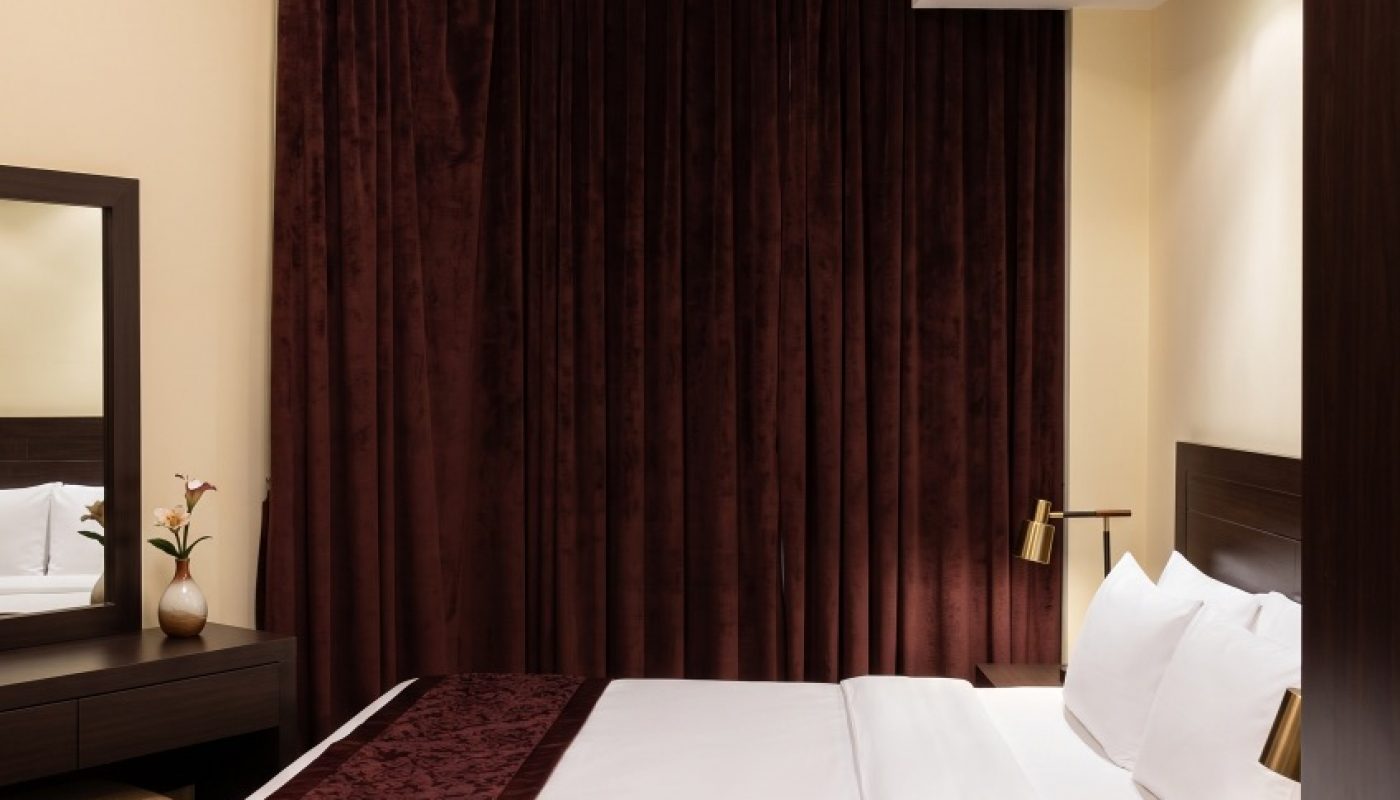 A hotel room with a double bed, white bedding, burgundy accents, a bedside table, a desk with a mirror, and dark burgundy curtains covering the window.