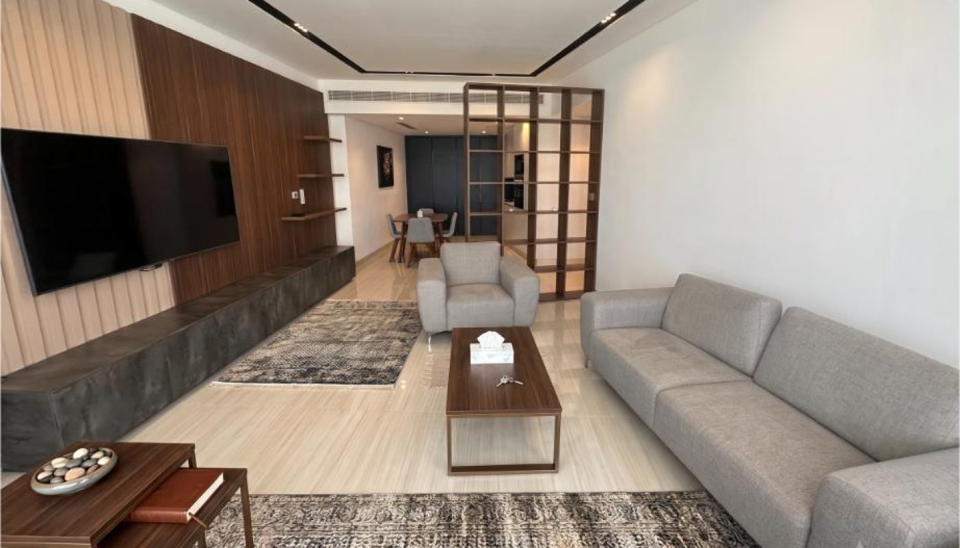 Modern living room with a gray sofa, coffee table, large TV on a wood-paneled wall, and dining area visible in the background. Neutral colors and minimalistic decor throughout.