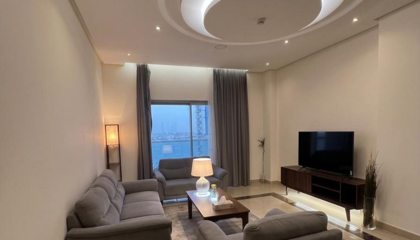 Modern living room with two gray sofas, a coffee table, TV on a stand, floor lamp, and large window with city view. Ceiling features a circular recessed light design.