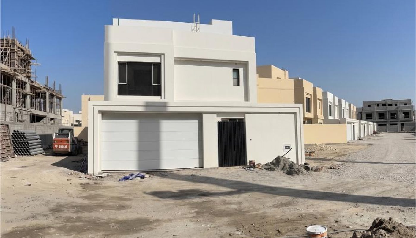 A modern two-story white house under construction, surrounded by unfinished ground and building materials, in a developing residential area.