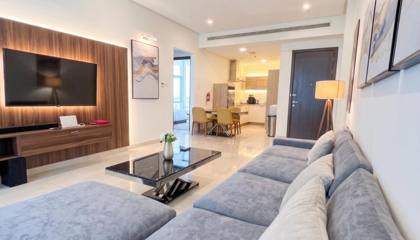 Modern living room with a large gray sectional sofa, wall-mounted TV, coffee table, and open view into a dining area and kitchen in the background.