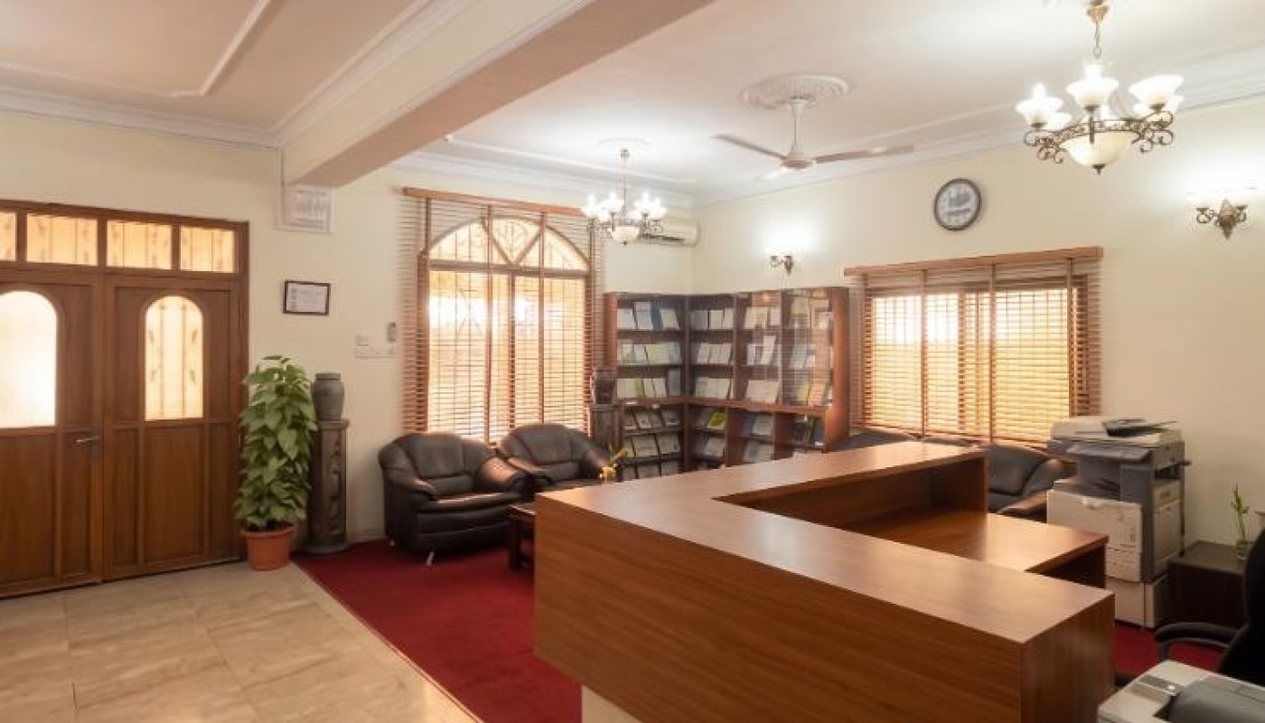 Reception area with a wooden desk, black chairs, bookshelves, indoor plant, photocopiers, and large windows with blinds. The floor is tiled and the ceiling has lights and ceiling fans.