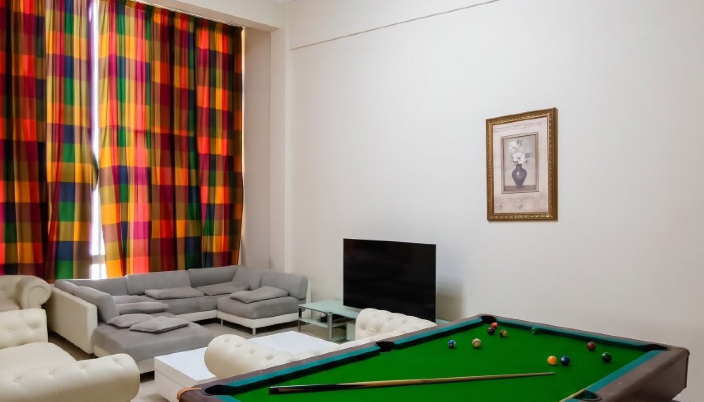 A room with a pool table, white sofas, a gray sectional, a TV, a coffee table, colorful plaid curtains, and a framed picture on a beige wall.