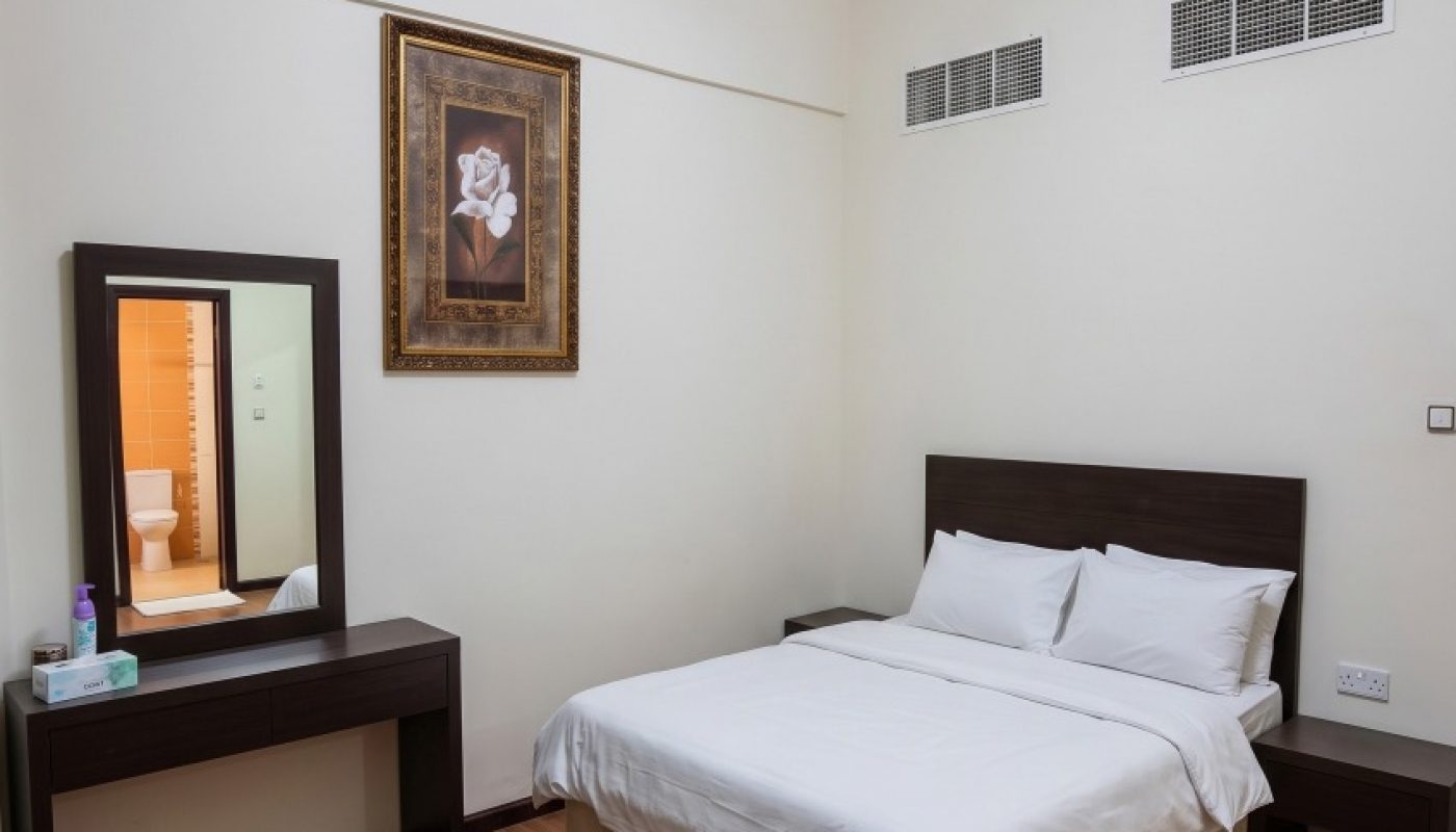 A neatly made bed with white linens beside a dark wood vanity and nightstand in a simple bedroom with a red and orange rug on a wooden floor.