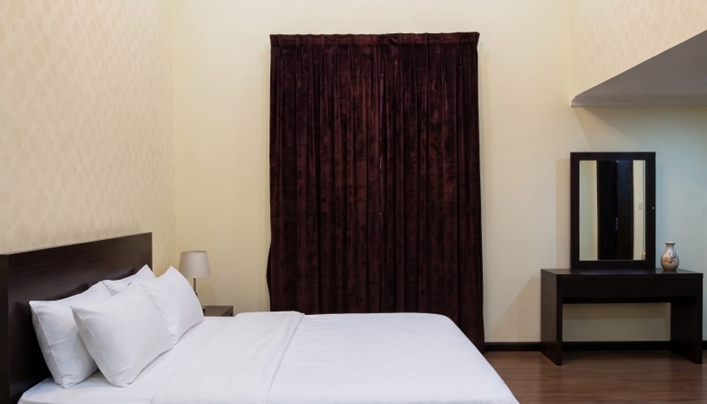 A neatly made bed with white linens sits against a wall, flanked by two nightstands. A curtained window and a vanity with a mirror are in the background.