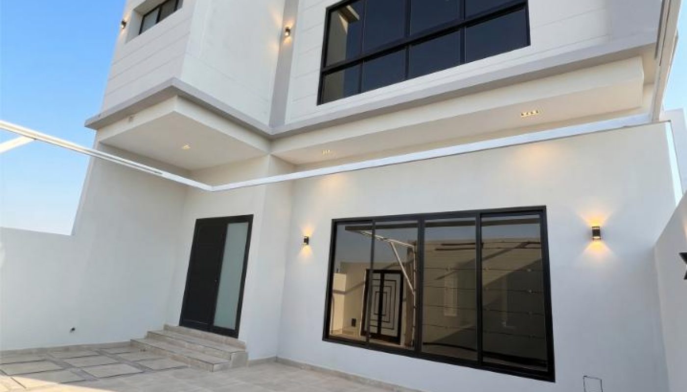 Modern two-story white house with large black-framed windows, exterior wall lights, tiled patio, and clear blue sky overhead.